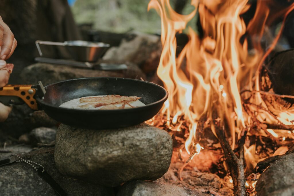 Skillet cooking pancakes and bacon over an open campfire