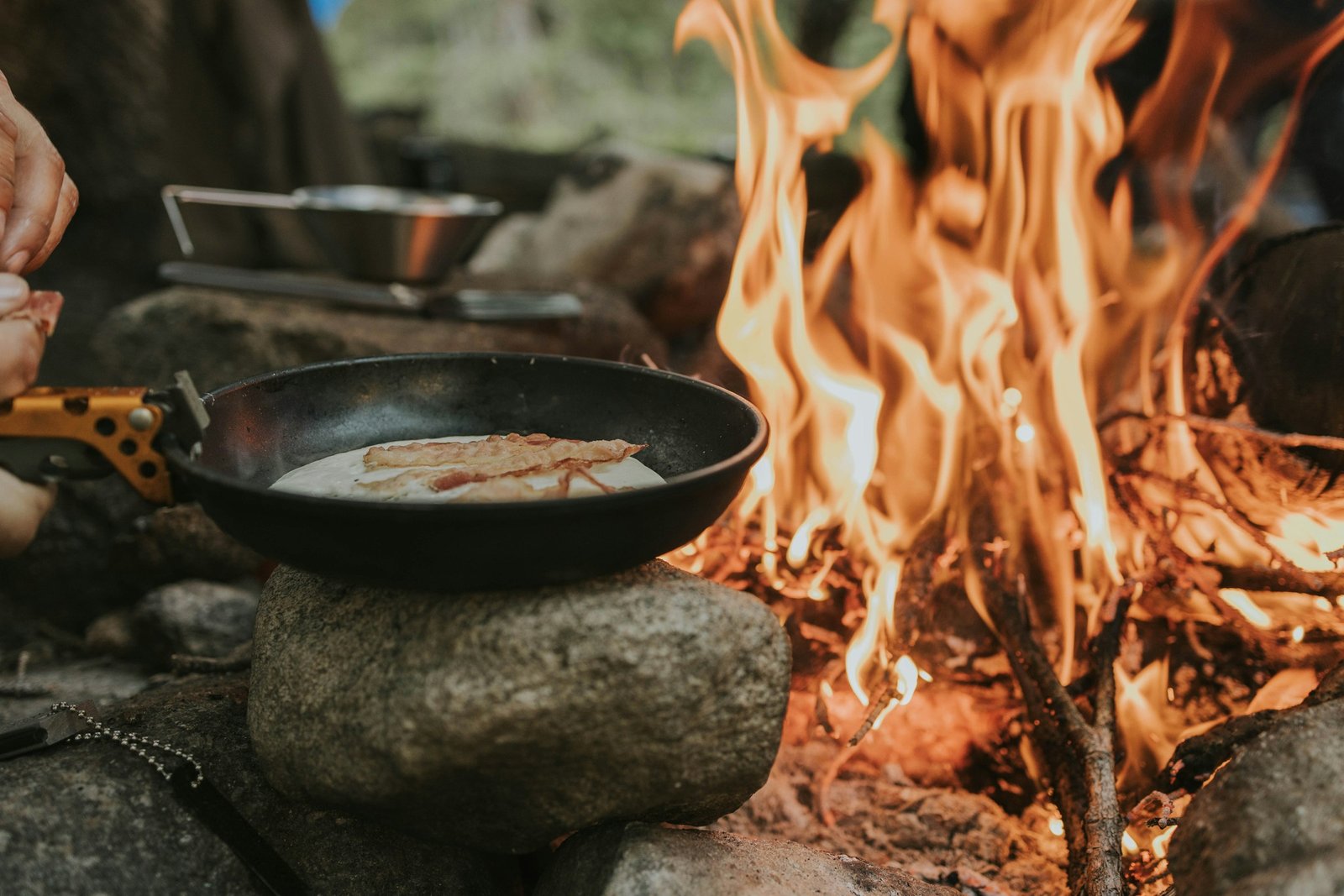 Skillet cooking pancakes and bacon over an open campfire